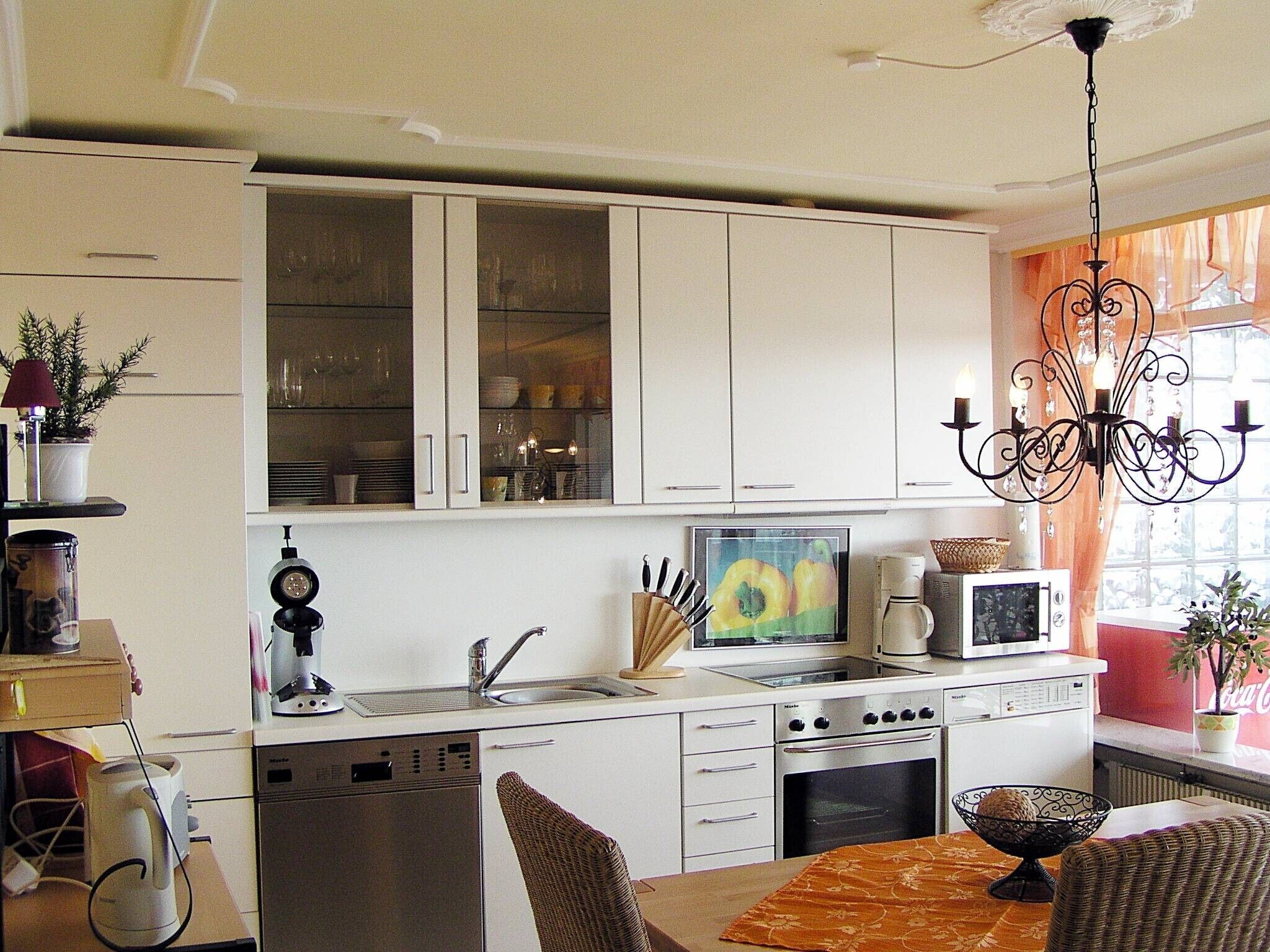 A compact kitchen with white cabinets, a dishwasher, an oven, and a microwave. There is a dining table with chairs, a chandelier, and a window with curtains.