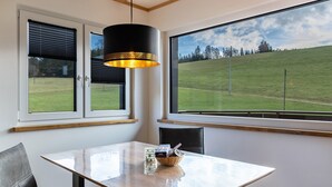 A dining area with a table, chairs, a hanging light fixture, and a large window with a view of a grassy hill. - Apartment 'Bergpanorama' with Mountain View, Shared Terrace and Wi-Fi (Triberg im Schwarzwald)