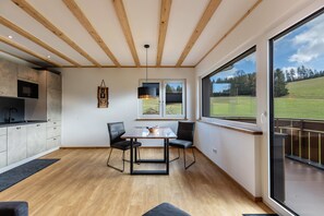 A modern kitchen with a dining area, wooden beams, and a large window with a view of a grassy field.