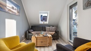 Un espacio habitable tipo loft con techo inclinado, un sofá gris, un sillón amarillo, una mesa de centro de madera y una ventana pequeña.