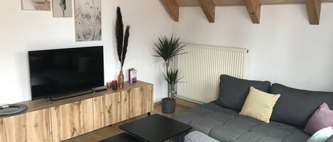 Een moderne woonkamer met een grote grijze bank, een flatscreen televisie, een houten salontafel en een plafond met zichtbare balken.
