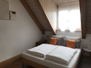 Une chambre avec un plafond en pente, un lit recouvert de draps blancs et une fenêtre ornée de rideaux légers.