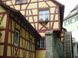 Un bâtiment à colombages avec une cheminée en pierre, une fenêtre agrémentée d’une jardinière et une porte bien visible.