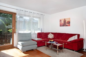 Un salon avec un canapé rouge, un fauteuil gris, une table basse en verre et un tableau accroché au mur.