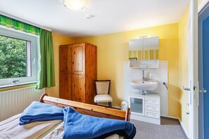 Une chambre à coucher comprenant un lit, une armoire en bois, une table de chevet blanche, un miroir et une fenêtre avec des rideaux.