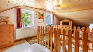Quarto com teto de madeira, janela com cortinas, radiador, cômoda e uma pequena casa de brincar de madeira.