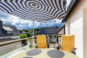 Un balcon agrémenté d’un parasol à rayures, de deux chaises jaunes à motifs et d’une table dont le plateau est en verre.