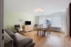 Una sala de estar con sofá, una mesa de comedor con sillas, televisor y ventana con cortinas.