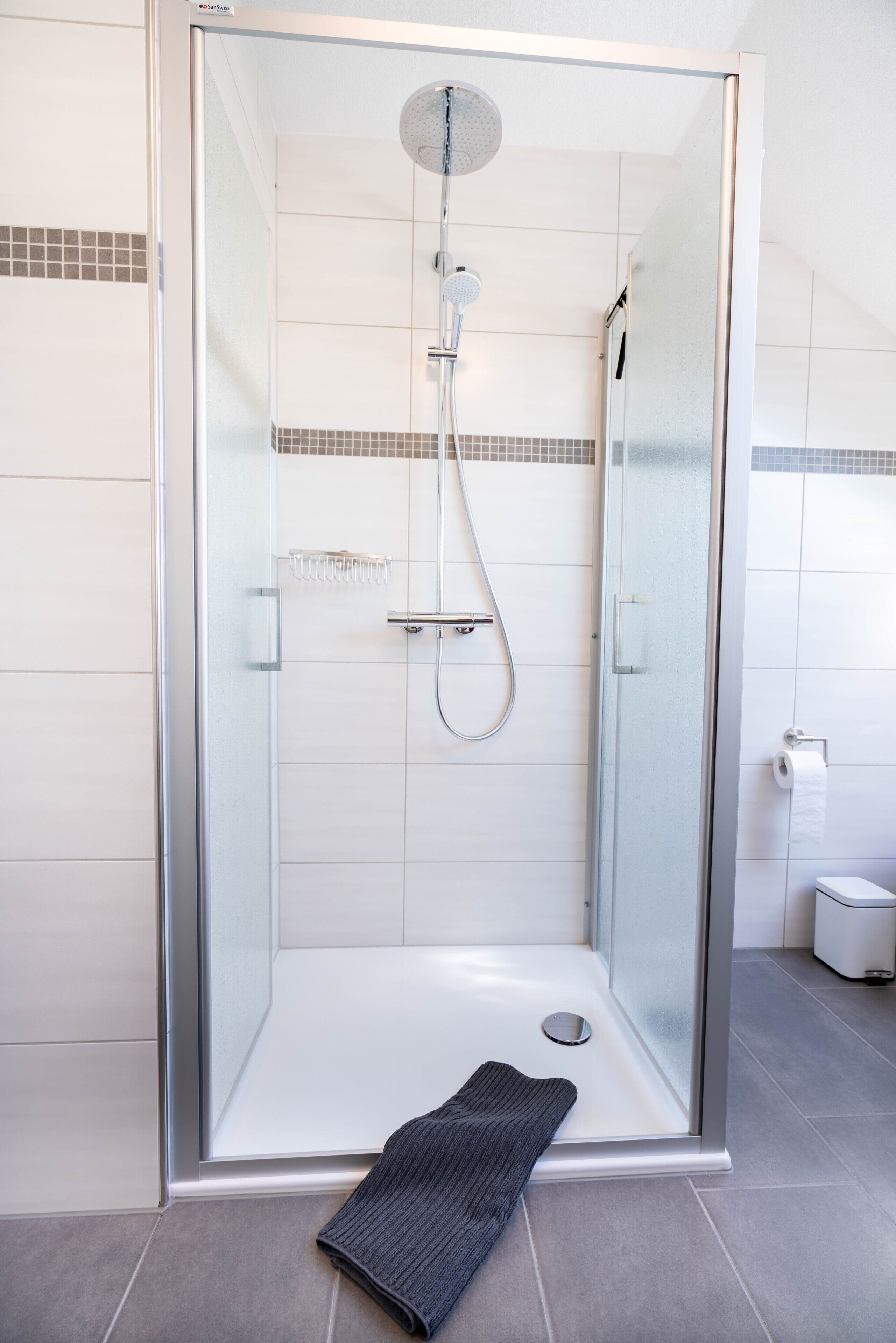 En dusch med glasväggar runt om, ett duschmunstycke som kan hållas i handen, en glasdörr och en handduk på golvet.