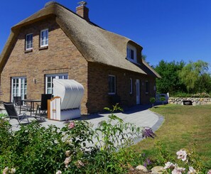 A brick house with a thatched roof, a patio with outdoor furniture, and a garden with flowers. - Apartment 'Käptn's Koje' with Private Terrace and Wi-Fi (Klanxbüll)