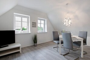 A modern dining area with a white table, grey chairs, a hanging light fixture, and a TV on a white stand. - Apartment 'Käptn's Koje' with Private Terrace and Wi-Fi (Klanxbüll)