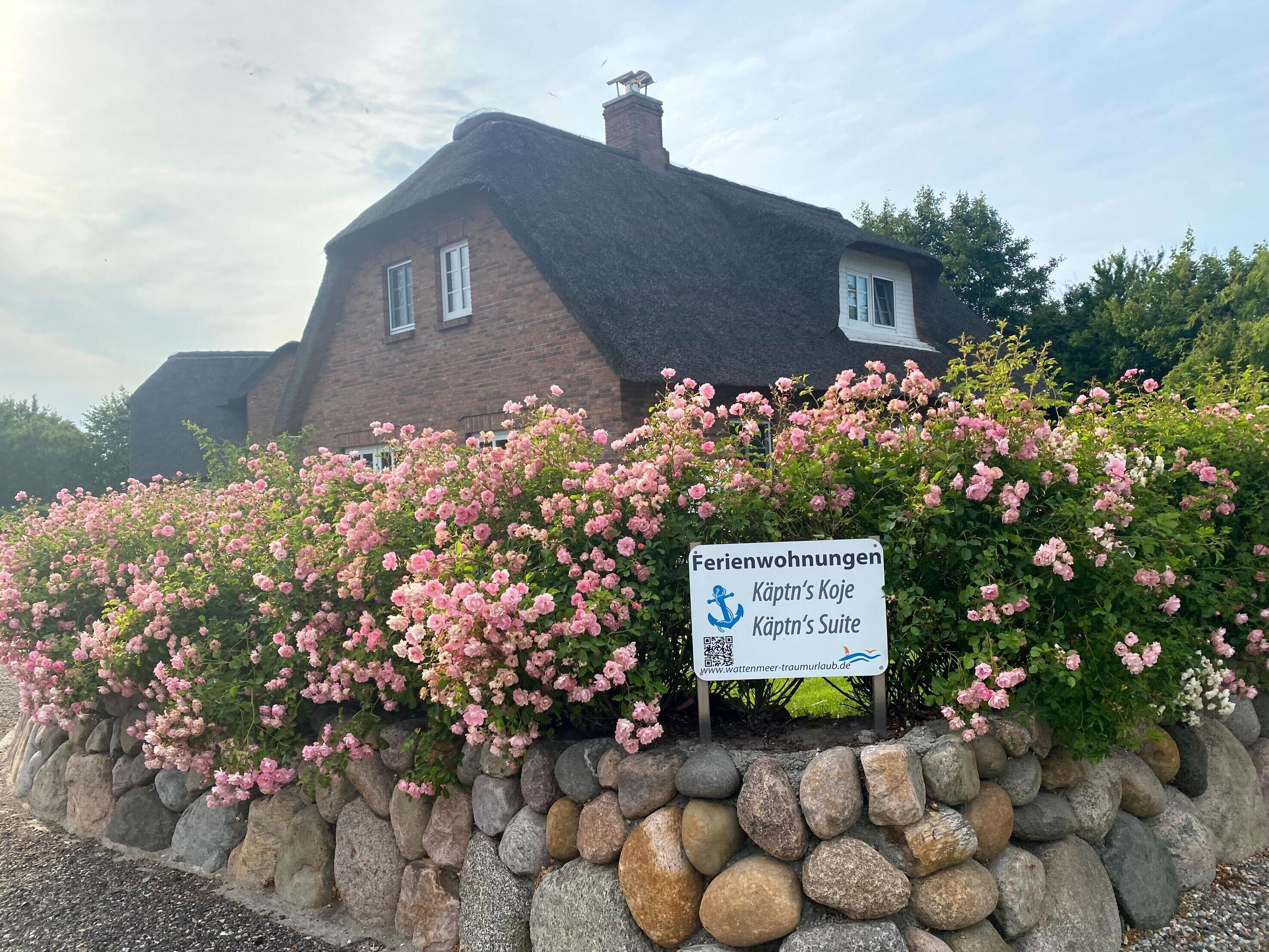 Une maison en briques avec un toit de chaume, entourée d’un mur de pierre et de buissons aux fleurs roses.