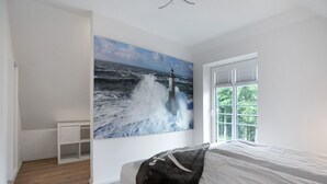 Une chambre avec un grand tableau représentant une vague océanique, un lit, une fenêtre avec des stores et une armoire blanche.