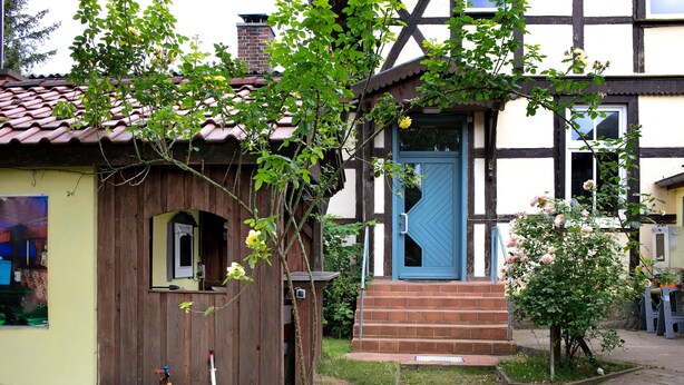 A traditional half-timbered house with a blue door, a wooden shed, and a garden with a small tree and flowers.