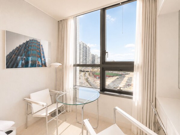 Ett modernt hotellrum med ett stort fönster, ett glasbord, vita stolar och en tavla som föreställer en byggnad.