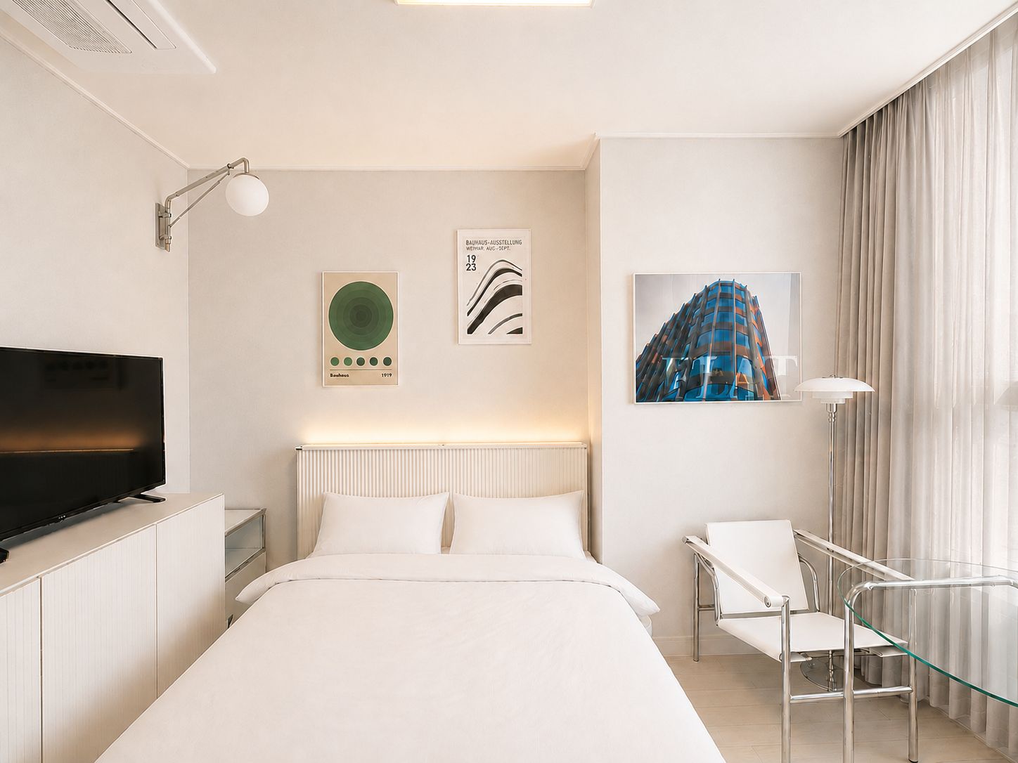 A modern hotel room with a bed, a flat-screen TV, a glass table, a chair, and artwork on the walls.