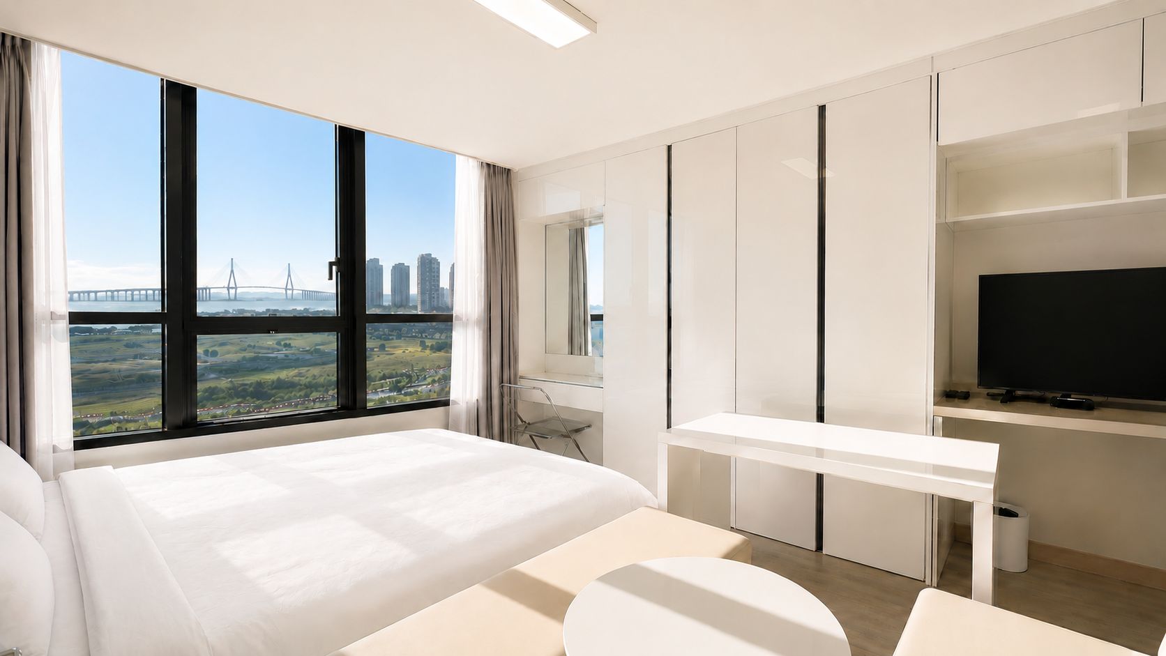 A modern hotel room with a large bed, a desk, a television, and a view of a cityscape through large windows.