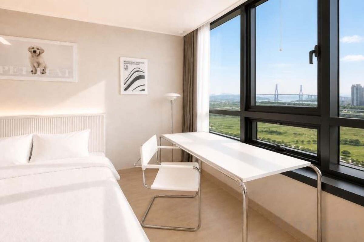 Habitación de hotel con cama, escritorio y un amplio ventanal con vistas a un puente y al paisaje urbano.