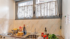 A kitchen with a stainless steel sink, a window with a leaf-patterned curtain, and various kitchen items like bottles and a cutting board. - Chiasso Apartments (Chiasso)