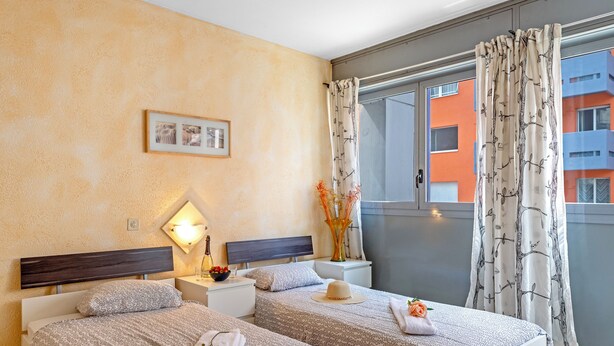 A hotel room with two beds, a nightstand with a lamp, a framed picture on the wall, and a view of buildings through the window. - Chiasso Apartments (Chiasso)