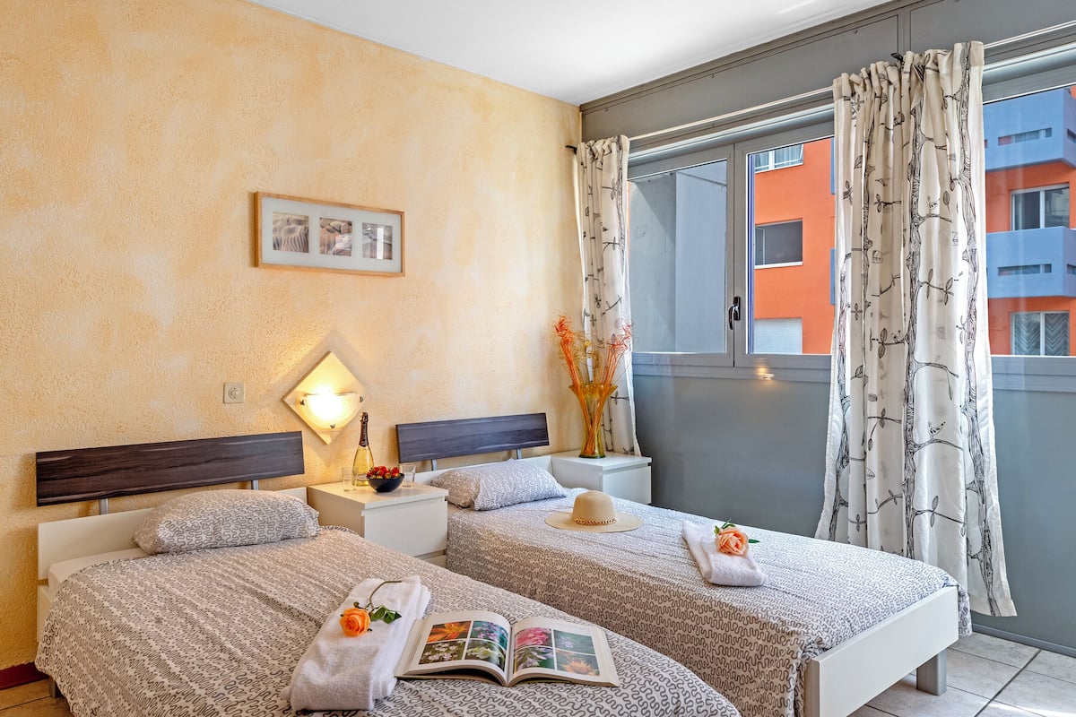 Une chambre d’hôtel avec deux lits, une table de chevet avec une lampe, un tableau encadré au mur et une vue sur les immeubles par la fenêtre.