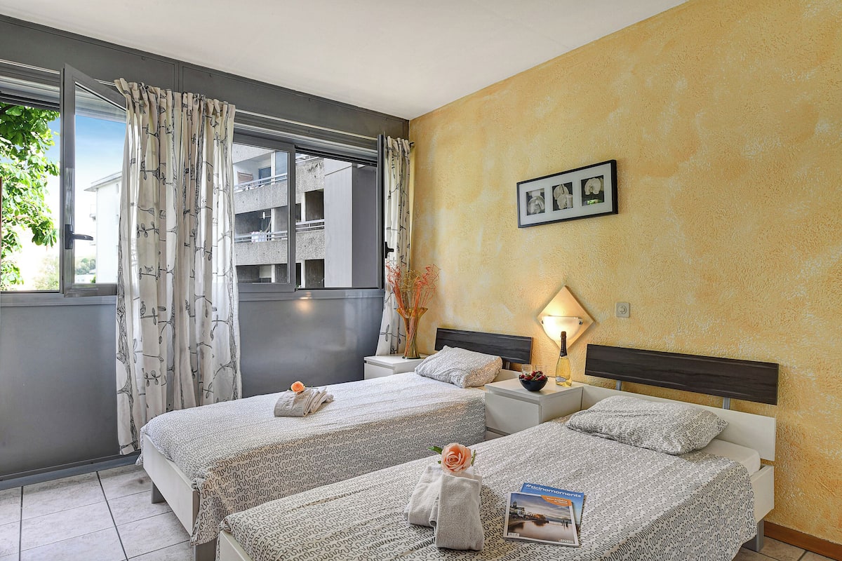 Une chambre d’hôtel avec deux lits, une fenêtre avec des rideaux, une table de chevet avec une lampe et un tableau encadré au mur.