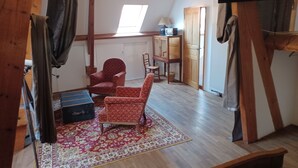 Une pièce avec un plafond en pente, des poutres en bois, un tapis à motifs, deux fauteuils à motifs, une petite table, un meuble et un radiateur.