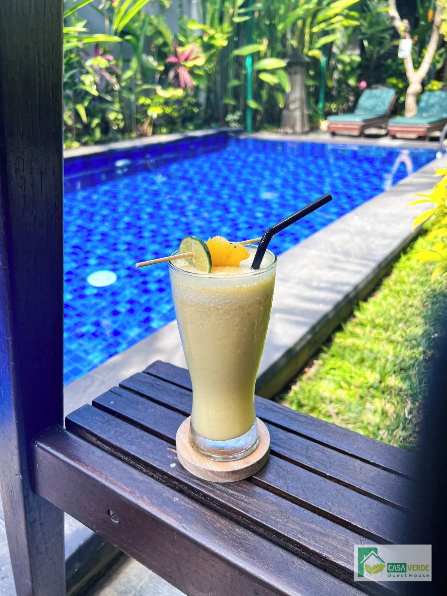 A glass of a light-colored beverage with a straw and a lime wedge on a wooden coaster by a poolside.