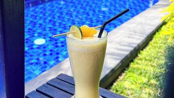 A glass of a light-colored beverage with a straw and a lime wedge on a wooden coaster by a poolside.