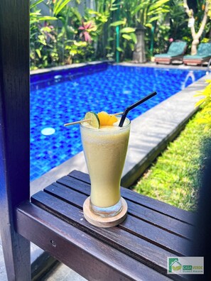 Un vaso de una bebida clara con pajita y una cuña de lima sobre un posavasos de madera junto a una piscina.