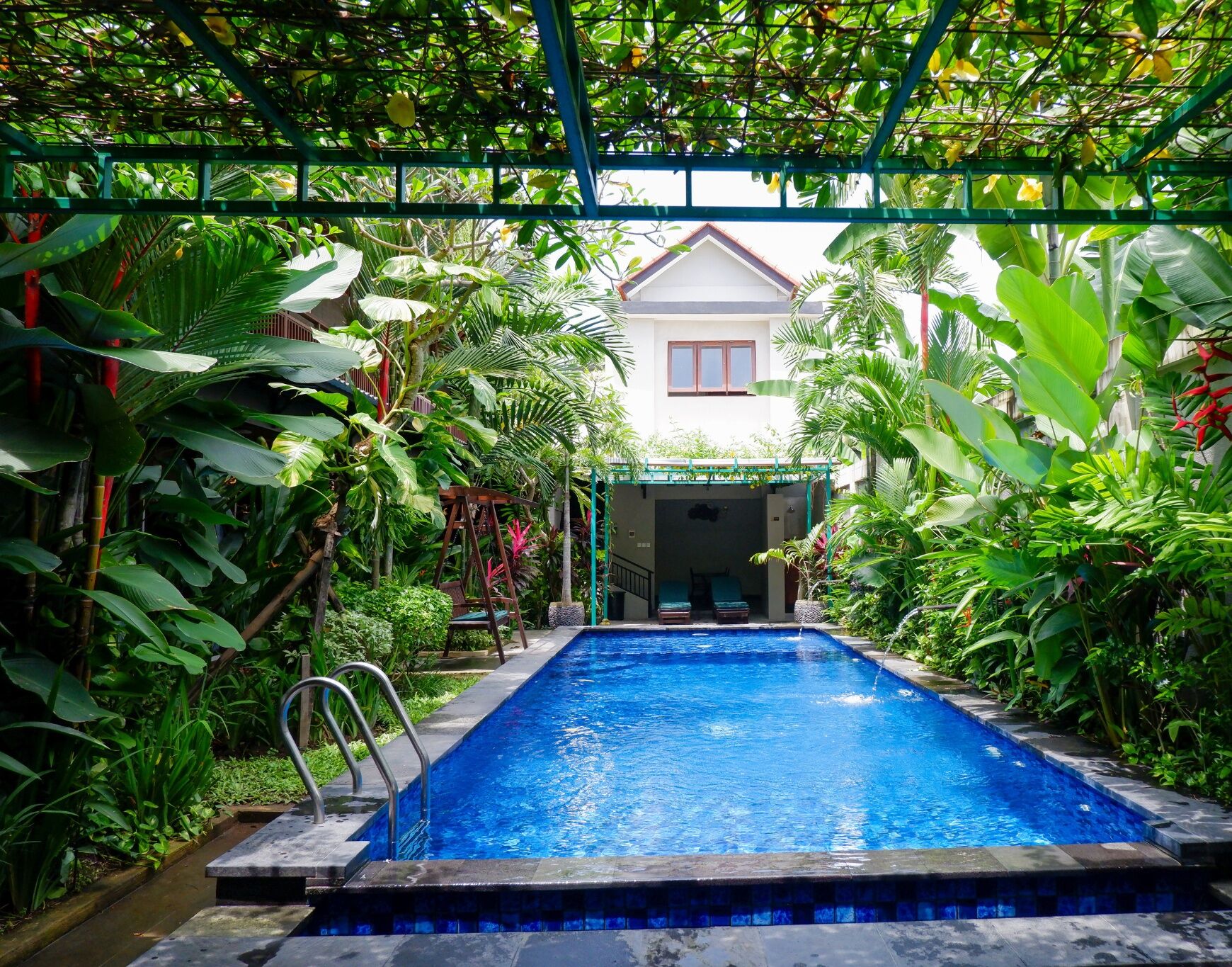 A rectangular swimming pool with a metal ladder, surrounded by lush tropical plants and a covered structure in the background.