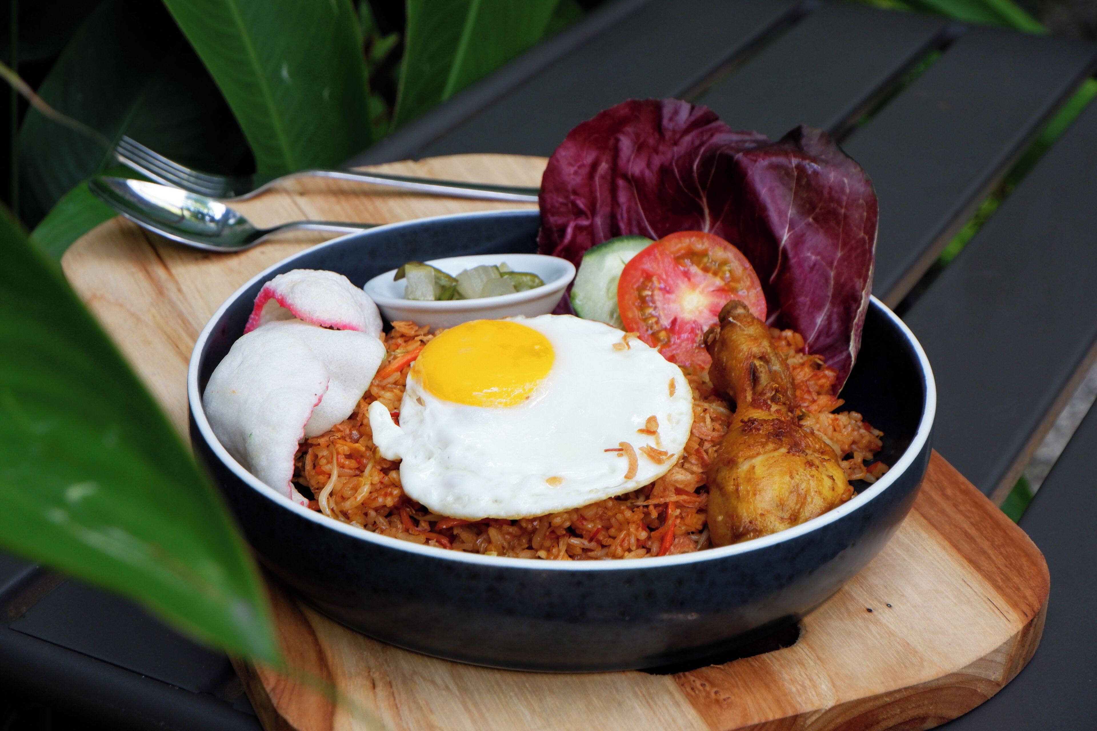 A bowl of food with a fried egg, vegetables, and a piece of meat on a wooden board.