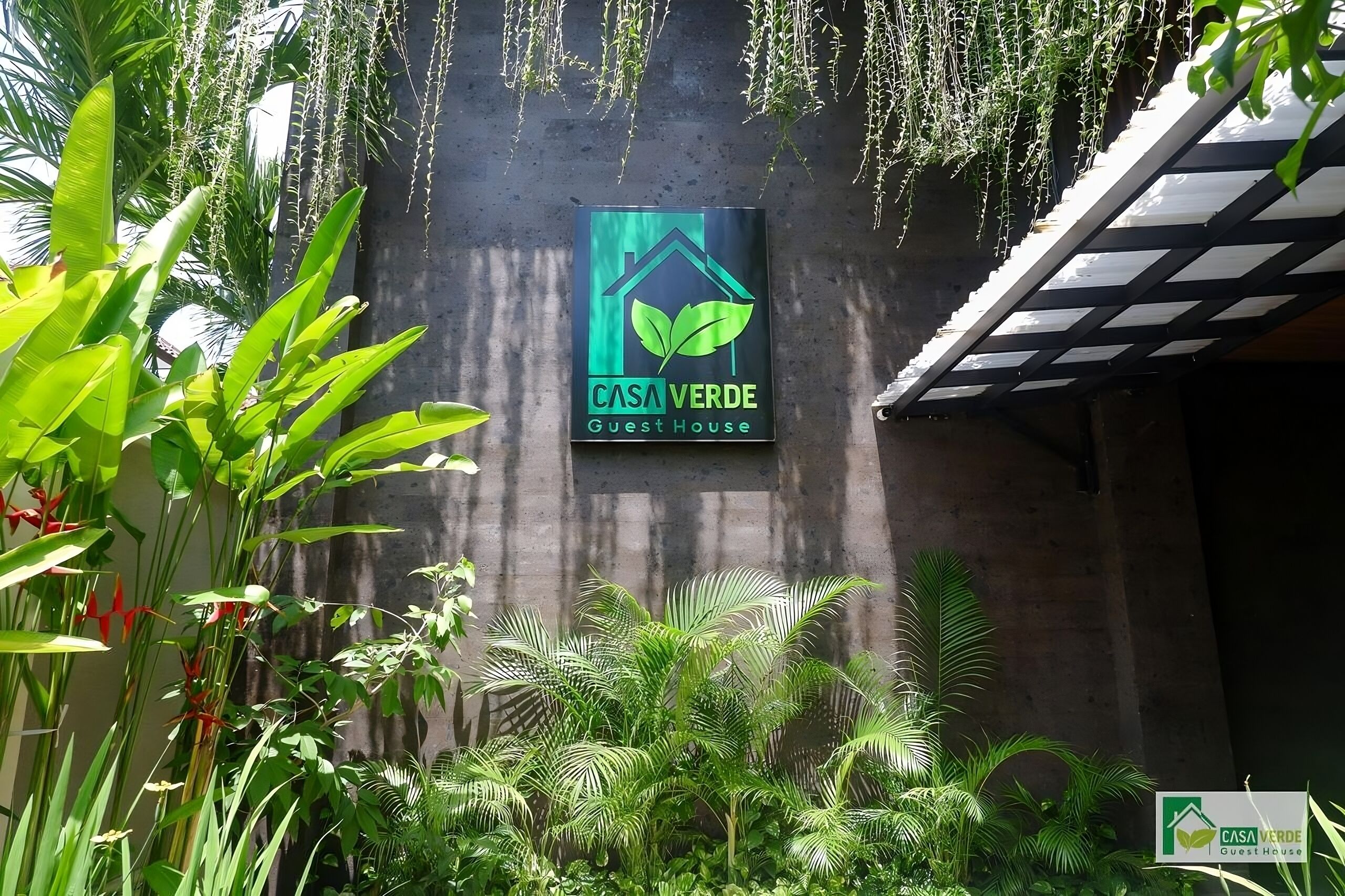 Een bord met de tekst 'CASA VERDE Guest House', met een logo van een groen blad, omringd door weelderige tropische planten.