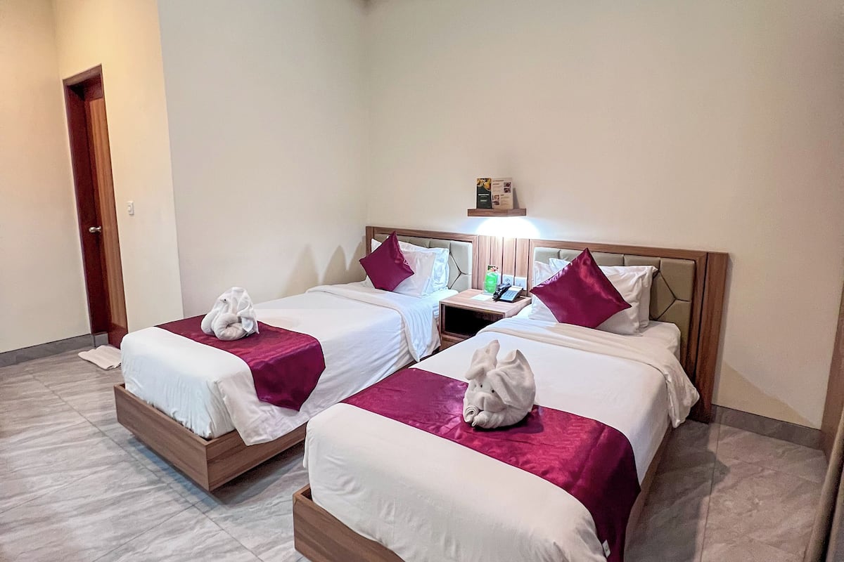 Quarto de hotel com duas camas, cada uma com estrutura de madeira e roupa de cama branca, piso de azulejos de mármore e uma pequena mesa de cabeceira com abajur.