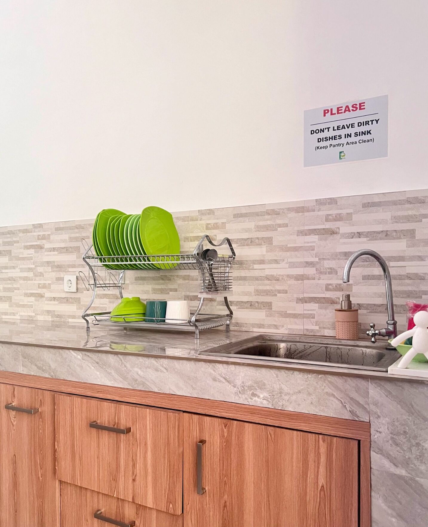 A kitchen with a dish rack holding green plates, a sink, and a sign requesting to keep the sink area clean.