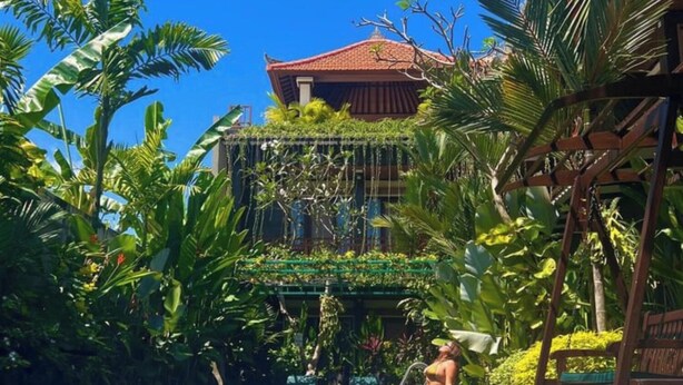 A pool with a tiled floor, surrounded by lush greenery and a building with a red roof in the background. - Casa Verde Guesthouse (Mengwi)