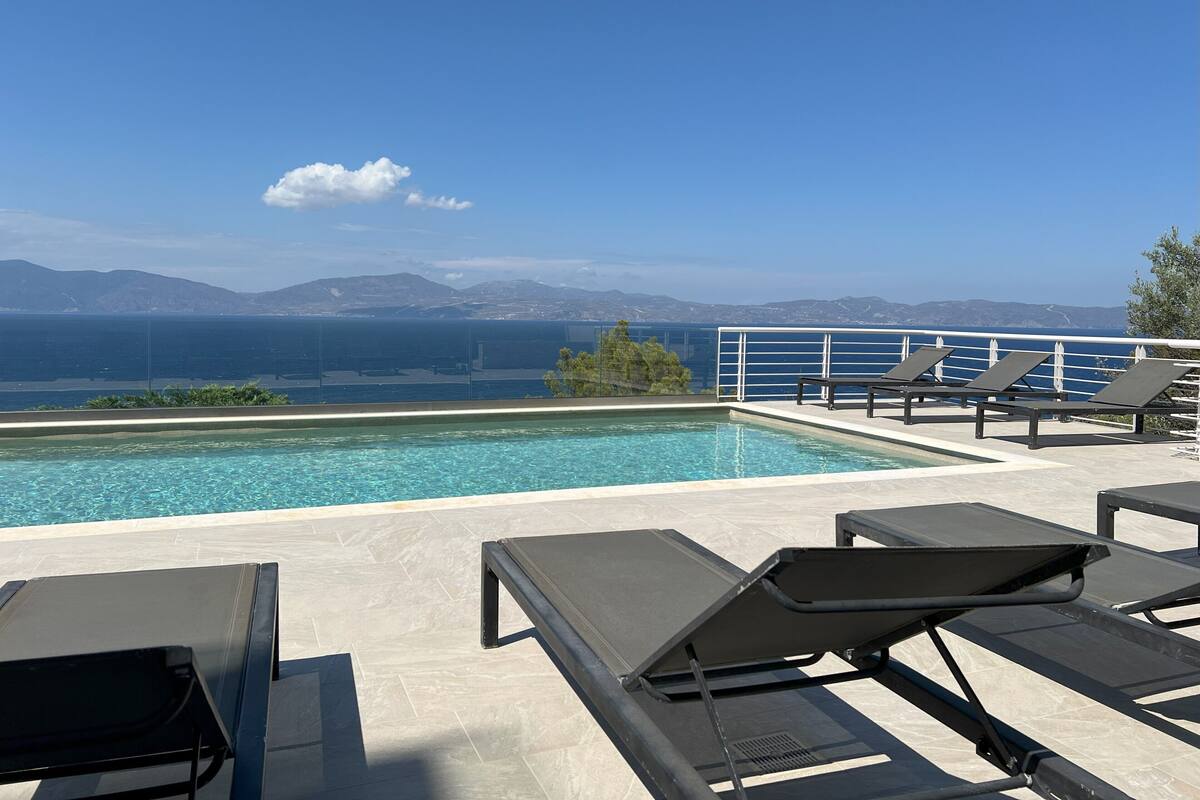 A rooftop pool with clear blue water, surrounded by lounge chairs and a view of the sea and mountains.