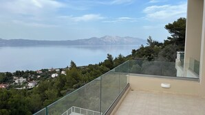 A balcony with a glass railing overlooking a lake, a pool, and a view of a town and mountains in the distance.