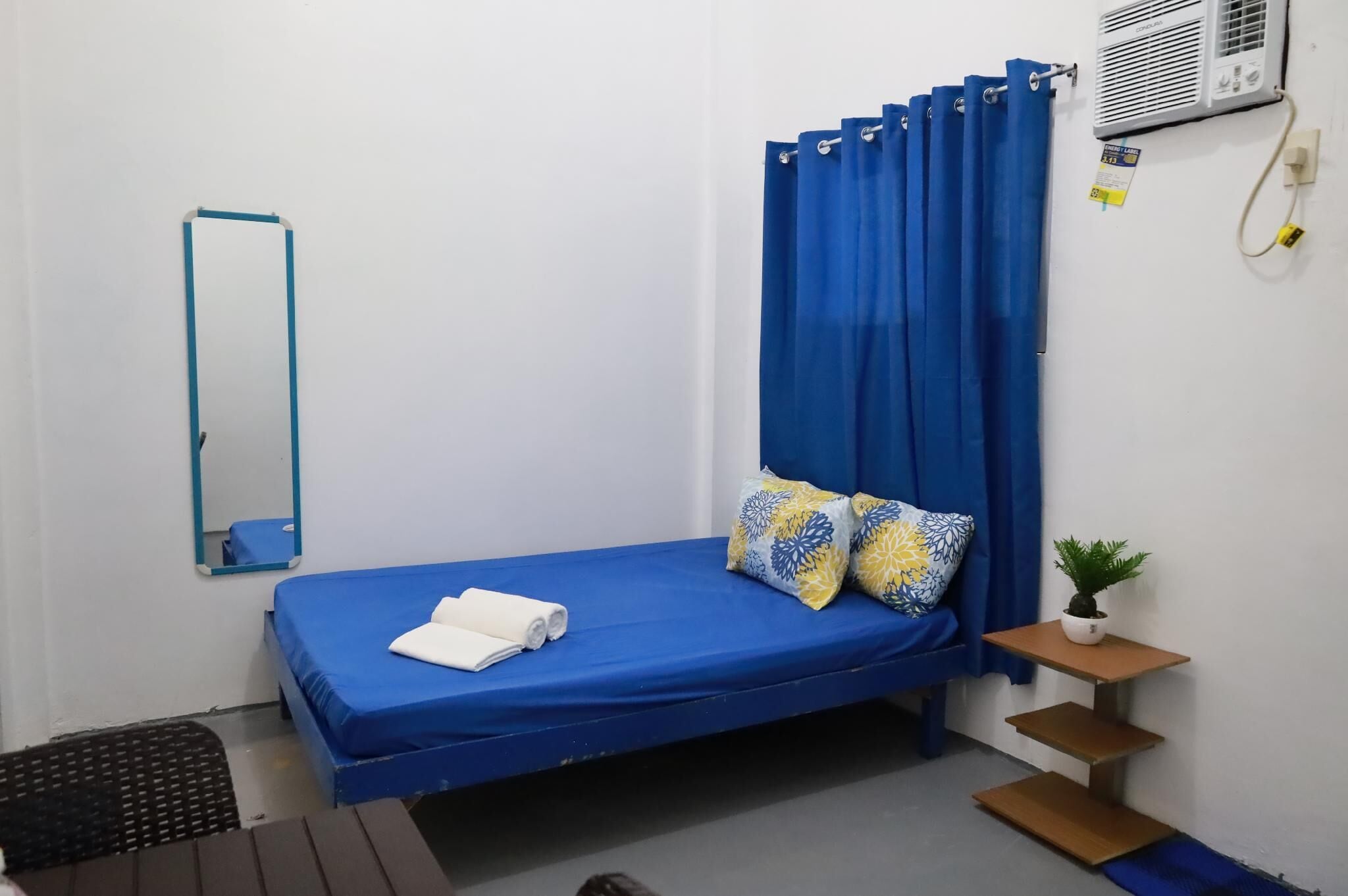 A small, neatly arranged bedroom with a single bed, a mirror, a small table with a plant, and a wall-mounted air conditioner.