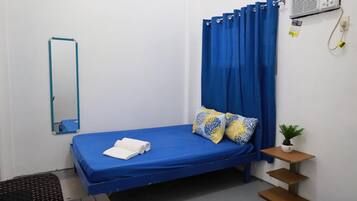 A small, neatly arranged bedroom with a single bed, a mirror, a small table with a plant, and a wall-mounted air conditioner.