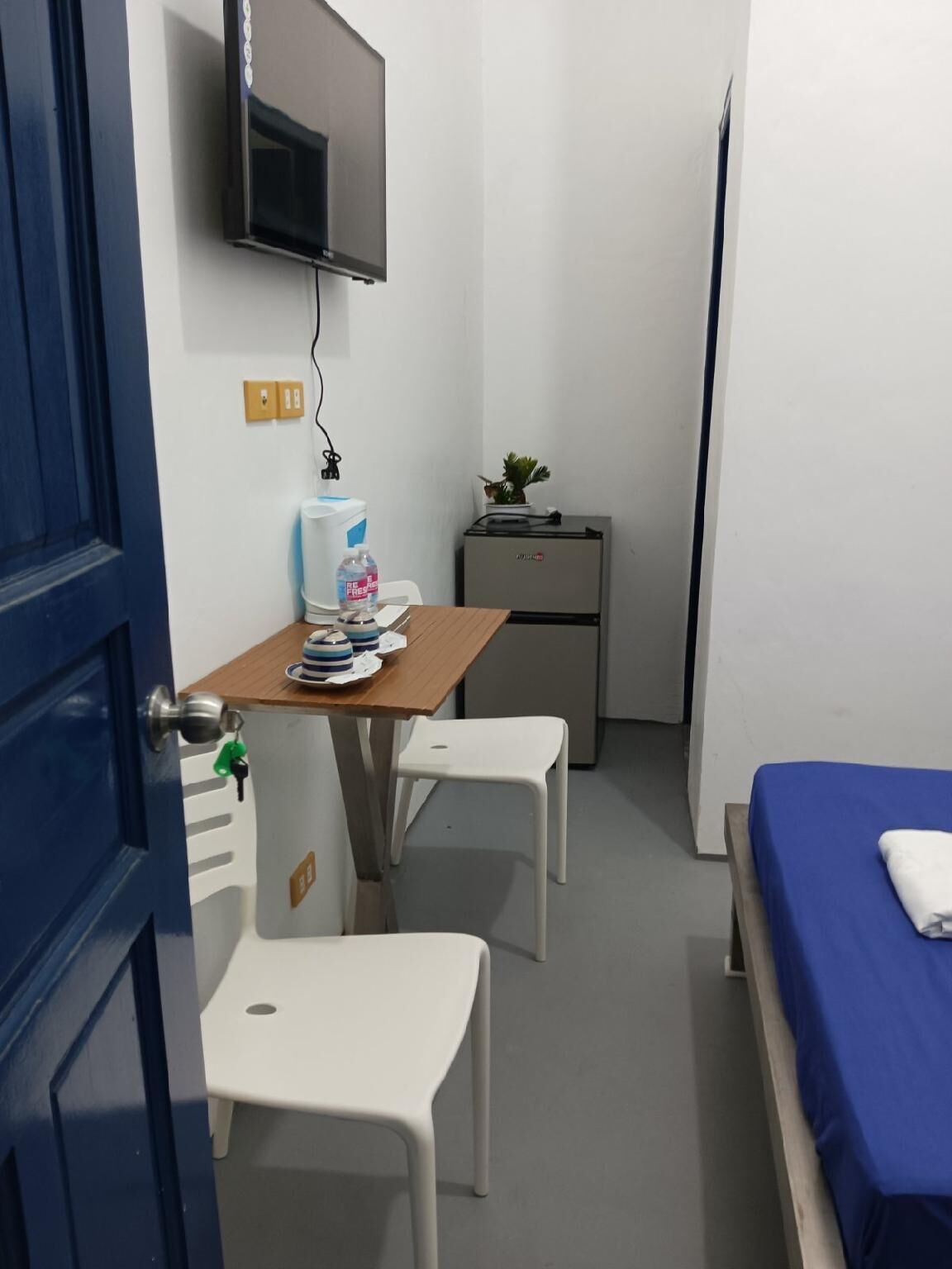 A small, compact room with a bed, a desk with a chair, a small refrigerator, a television mounted on the wall, and a door with a key lock.