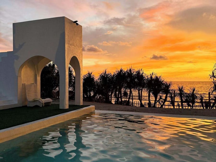 Zona de piscina con una estructura arquitectónica moderna que presenta arcos, vista a la playa y palmeras.