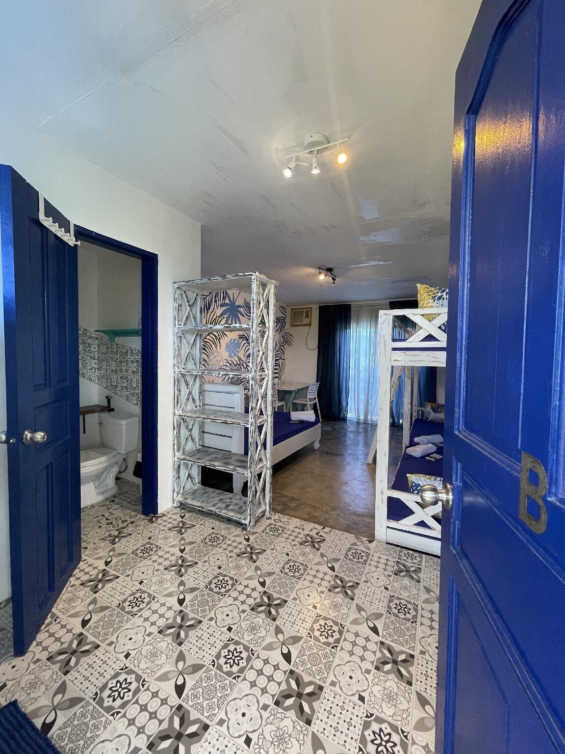 A room with a patterned floor, a bunk bed, a white shelf unit, and a blue door.