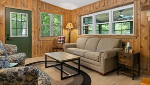 A cozy room with wooden walls, a green door, and large windows. There is a beige sofa, a patterned armchair, and a small table with a lamp. - Heron Hideout: Shenandoah Riverfront Retreat! (Luray)