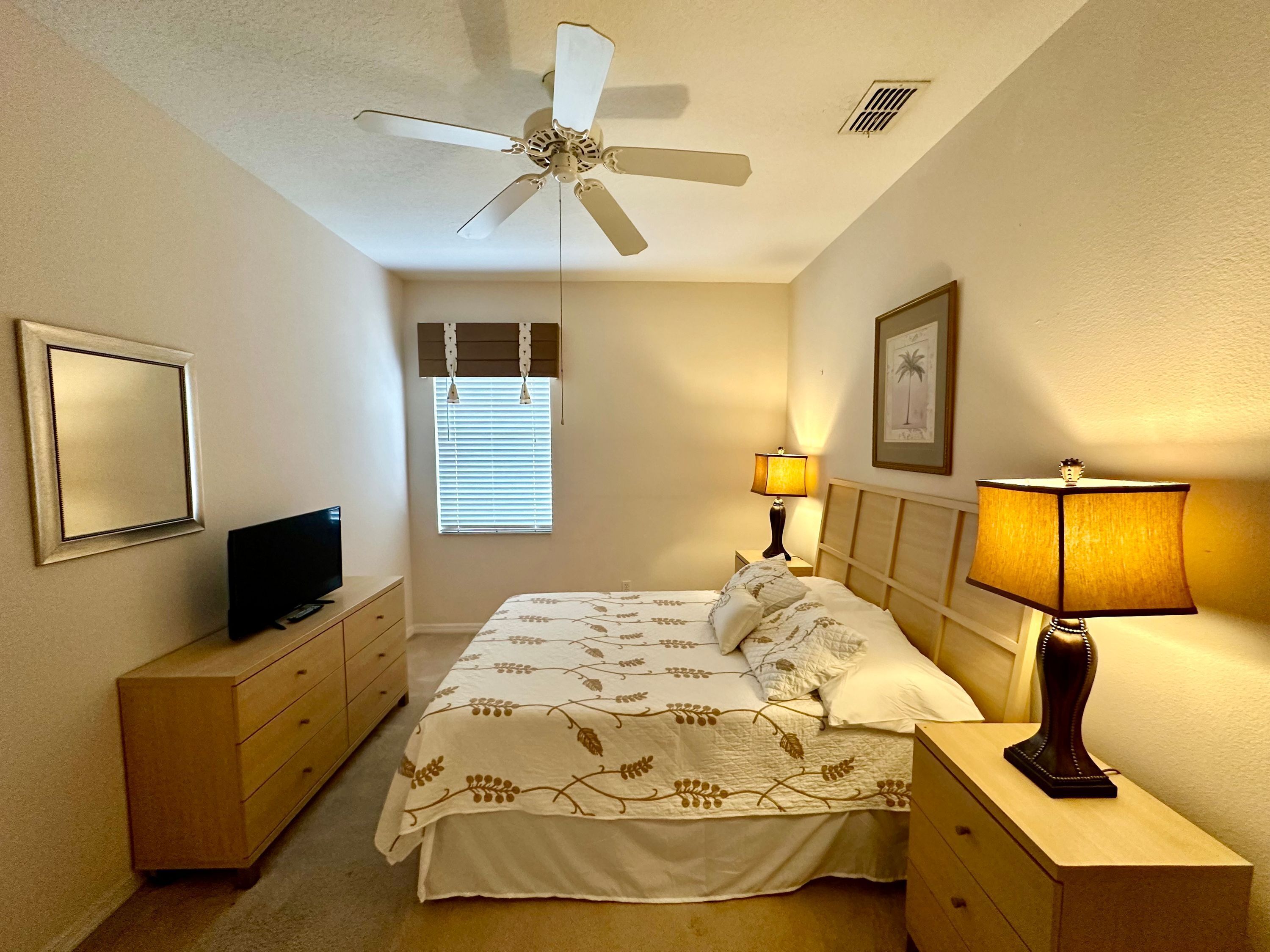 Une chambre d’hôtel équipée d’un lit, de lampes de chevet, d’un ventilateur de plafond, d’une télévision, d’une commode et d’une fenêtre avec des stores.