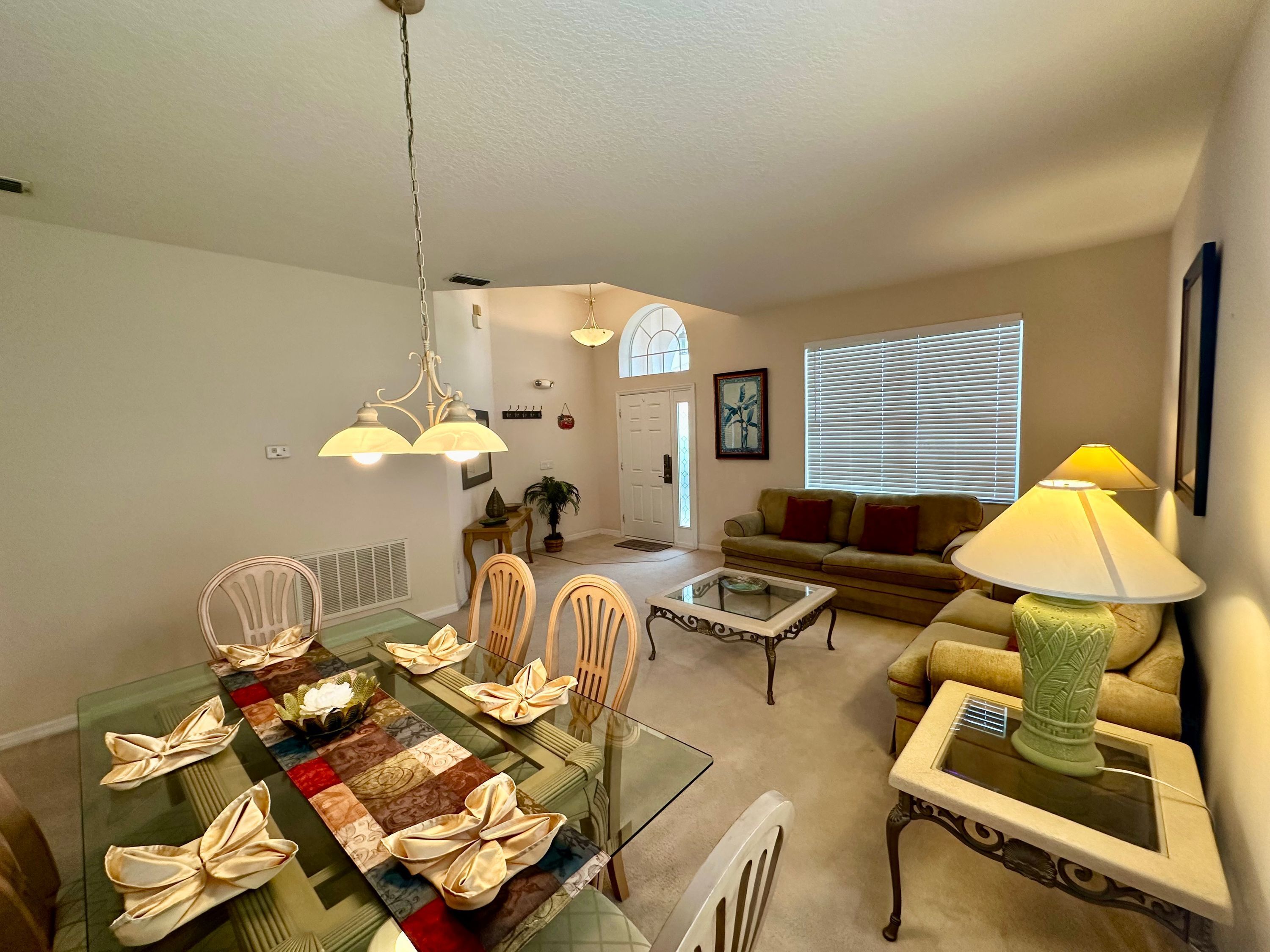 A dining area with a glass table, chairs, and a chandelier. There is a living room with a sofa and a lamp. The room has a door and a window with blinds.