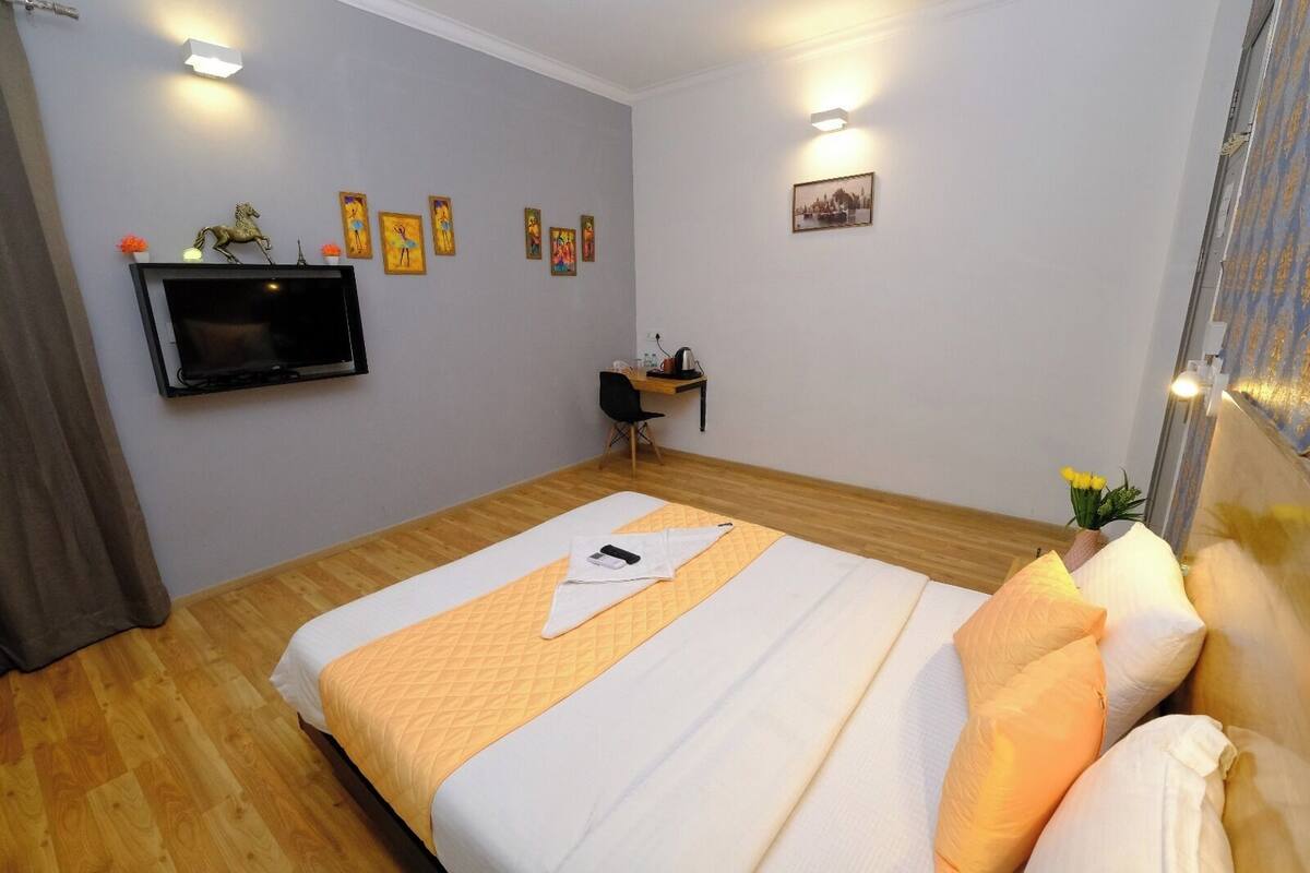 Quarto de hotel com uma cama, uma escrivaninha, uma cadeira, uma TV fixada na parede e algumas fotos emolduradas na parede.