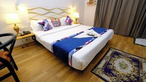 A hotel room with a bed, two bedside lamps, a nightstand, a chair, a rug, and a wall-mounted shelf with flowers. - Lime Tree Premier Near Medanta Medicity (Gurugram)