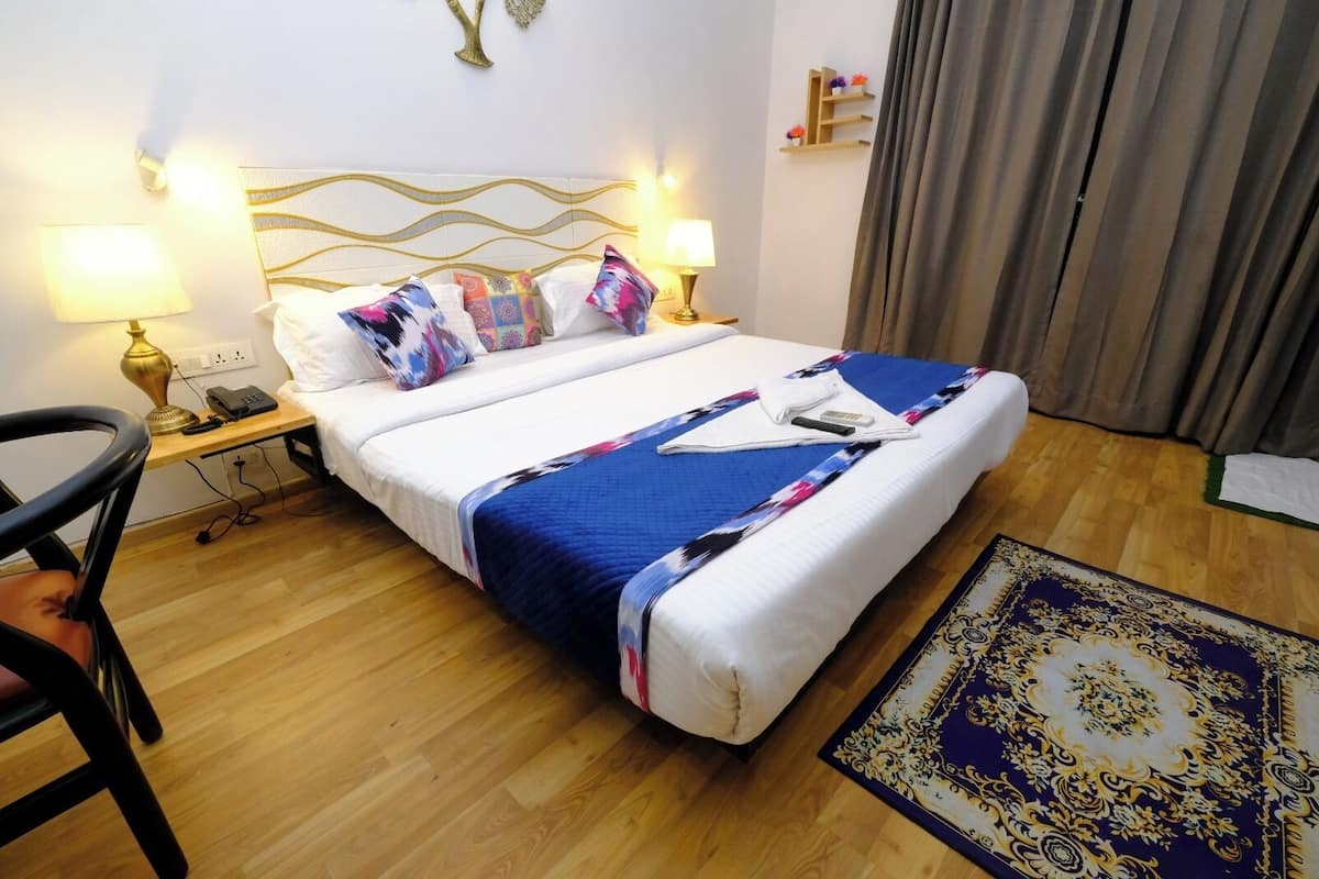Quarto de hotel com cama, duas luminárias de cabeceira, mesa de apoio, cadeira, tapete e prateleira na parede com flores.