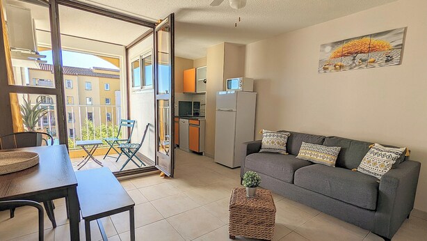 A living room with a grey sofa, a small table, a woven ottoman, and a balcony with a view of buildings and a plant. - Apartment close to port, beaches, golf course and ile des Loisirs with air conditioning (Agde)